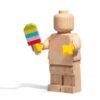 A premium lifestyle shot showing the solid oak Wooden Minifigure LEGO Originals model standing at 5 to 1 scale with yellow plastic hands holding built accessories